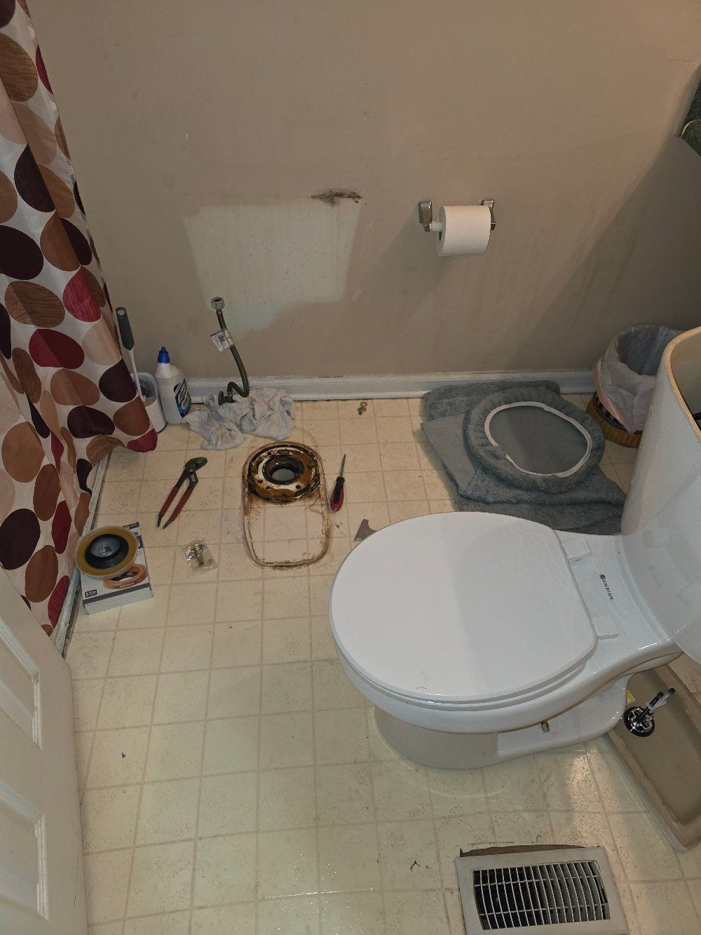 Small bathroom with toilet, sink, tiled floor, shower curtain, and cleaning supplies on the floor.