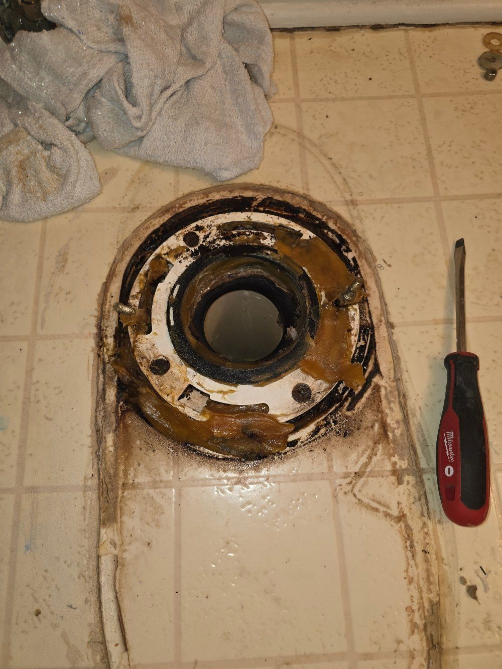 Dirty toilet flange on tile floor beside a red-handled screwdriver and a rag