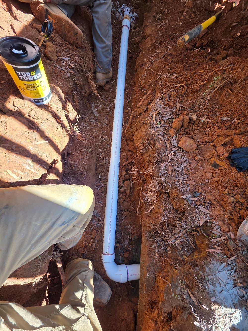 PVC pipe laid in a dirt trench beside a worker’s leg and a spray can of foam sealant