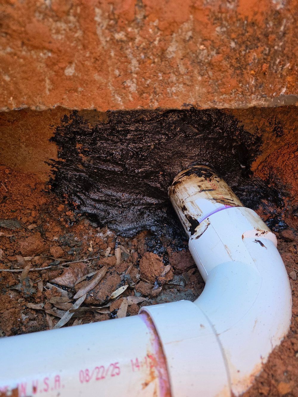 White PVC pipe elbow beside a dark damp patch against a brick wall and dirt ground