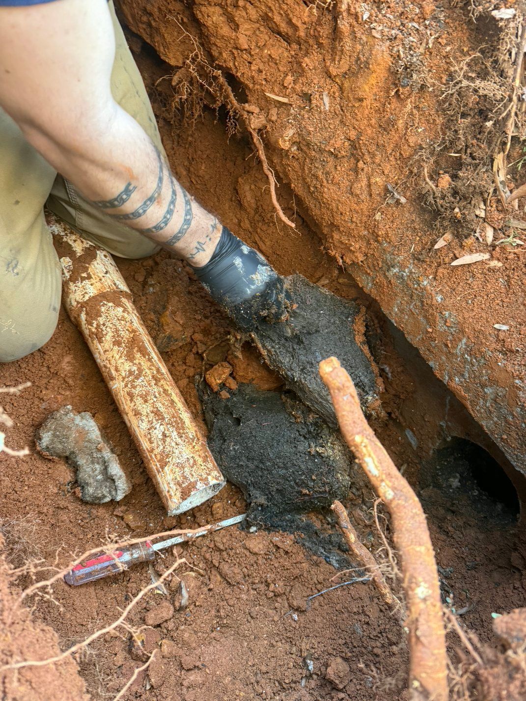 Person in a dirt trench handling a blackened pipe or root next to exposed orange piping and tools.