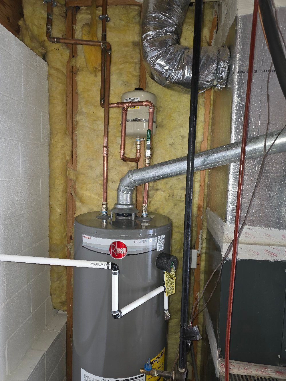 Water heater in a basement with exposed pipes and insulation, plus a large gray utility tank.