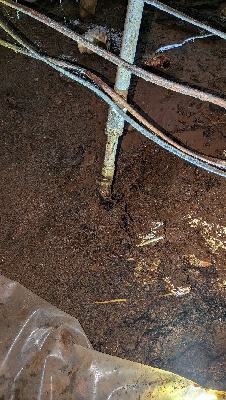 Rusty metal pipes crossing over dark soil or mud near a wall, with a plastic sheet at the bottom left
