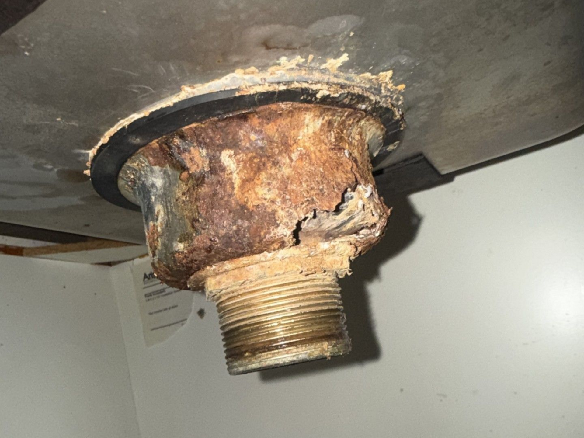 Rusty corroded metal pipe fitting under a tank or appliance, with heavy rust and damage.