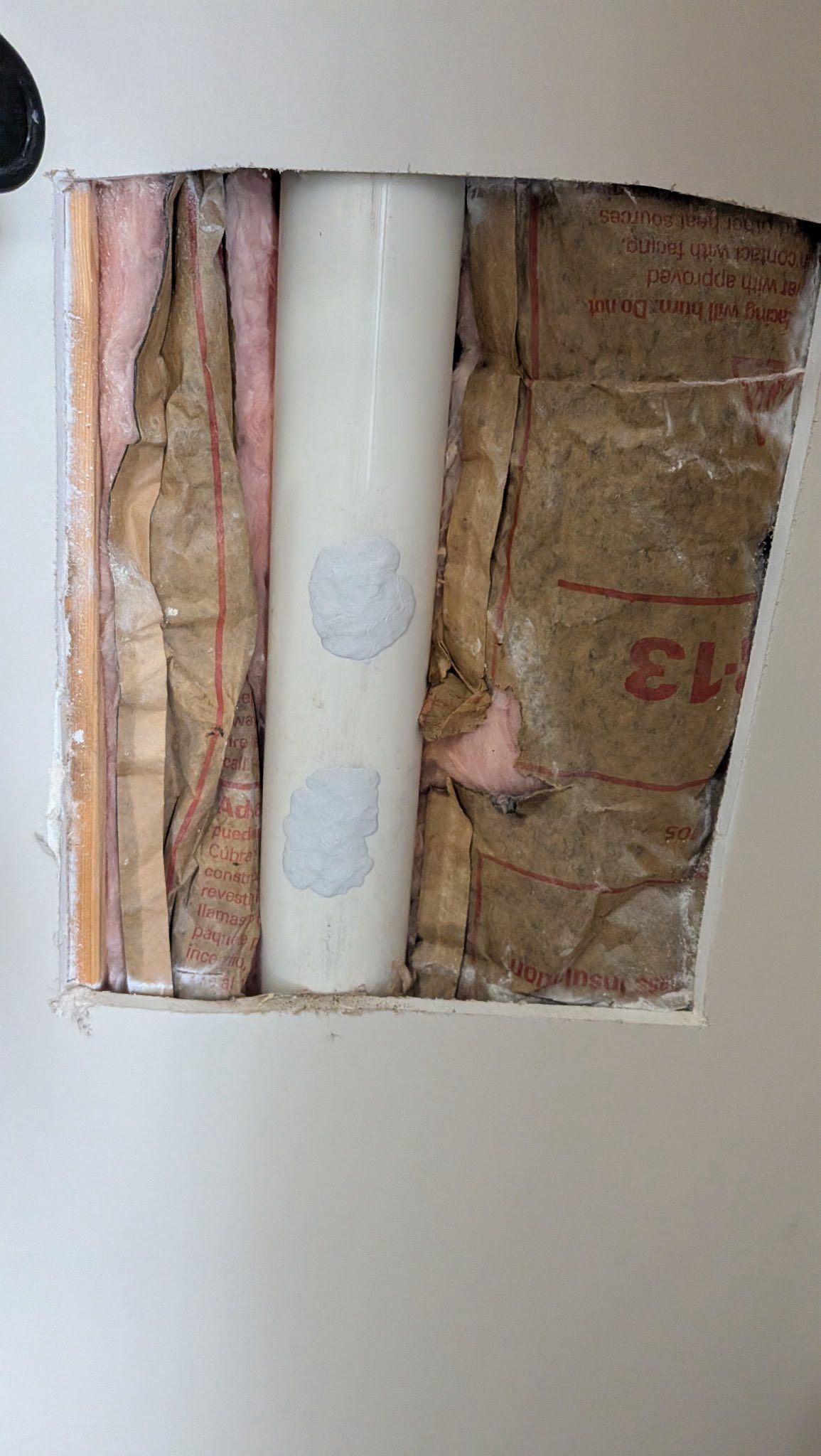 Wall cutout exposing insulation, a white pipe, and wooden framing inside.