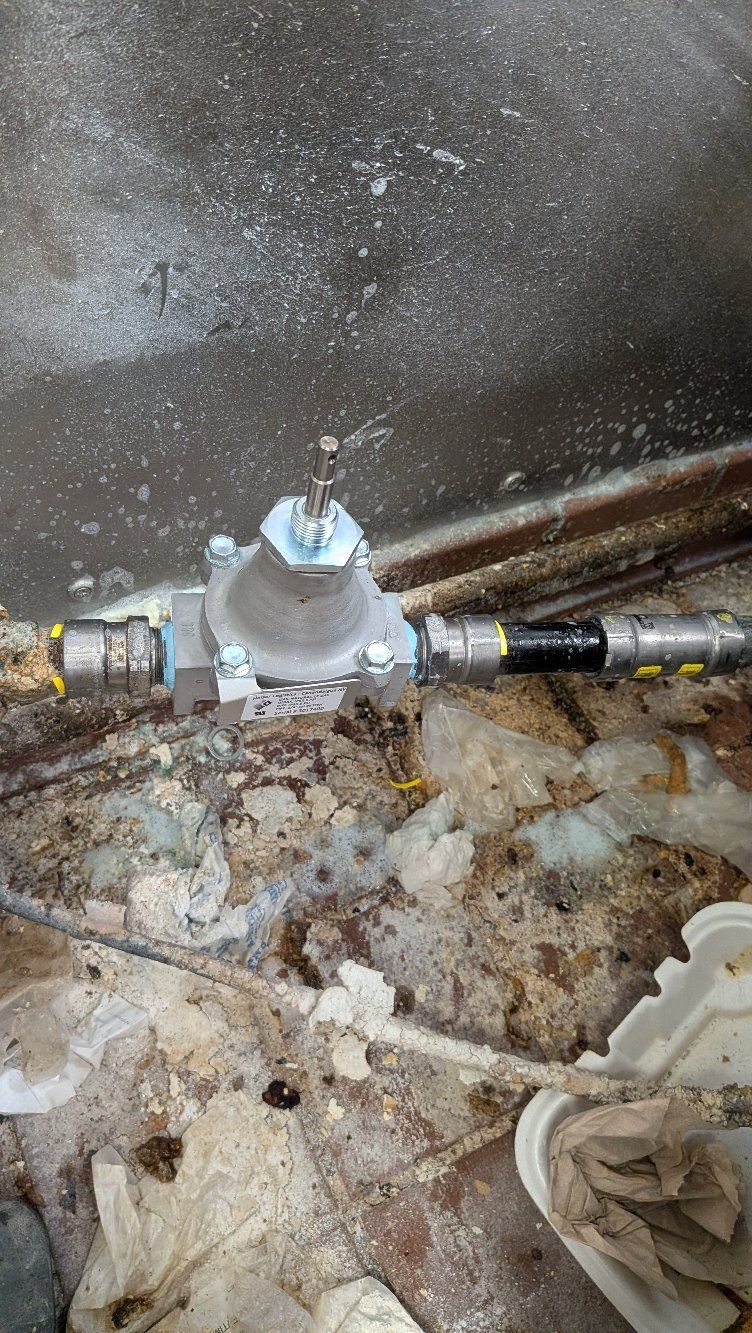 Corroded valve and pipes in a dirty, damp utility space with debris on the floor