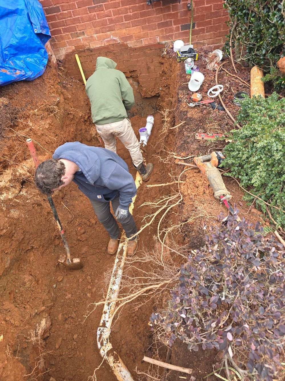Two people digging a trench beside a brick wall and plants, with tools and a white pipe in the dirt.