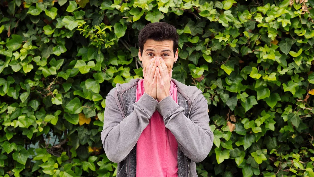 A man is covering his mouth with his hands in front of a green wall.