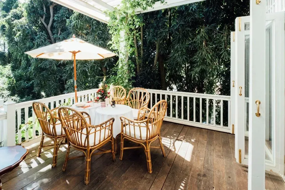 Wooden deck with a round table, bamboo chairs, and an umbrella, surrounded by greenery.