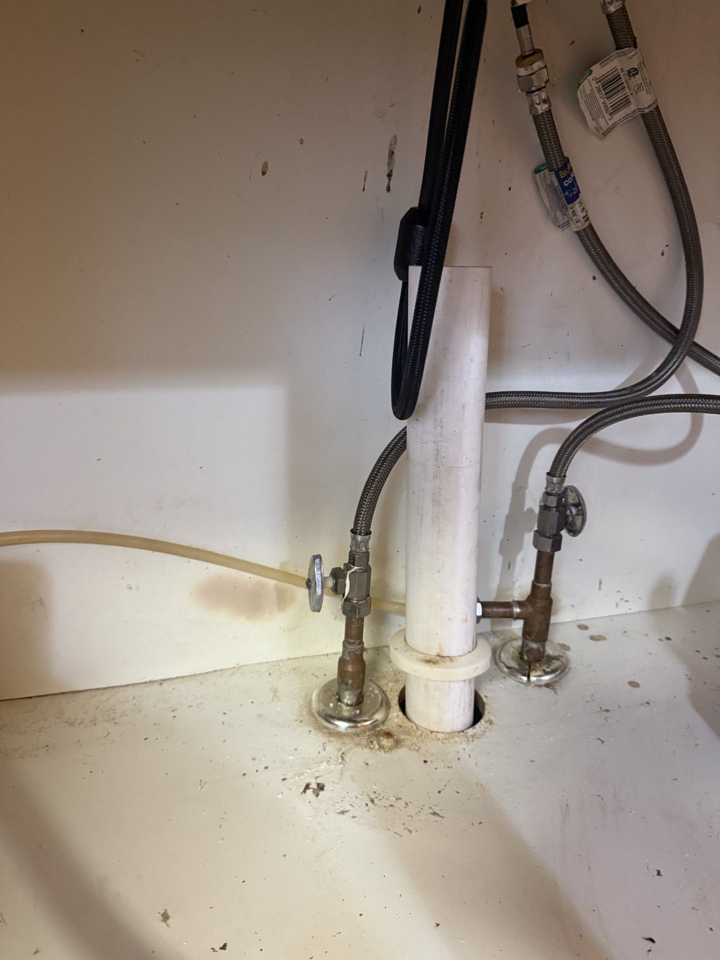 Under-sink plumbing with white pipes, hoses, and valves against a dusty cabinet wall