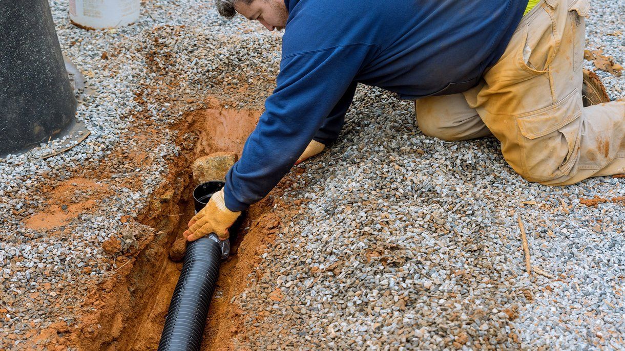 Man installing black drain pipe in a trench filled with gravel.
