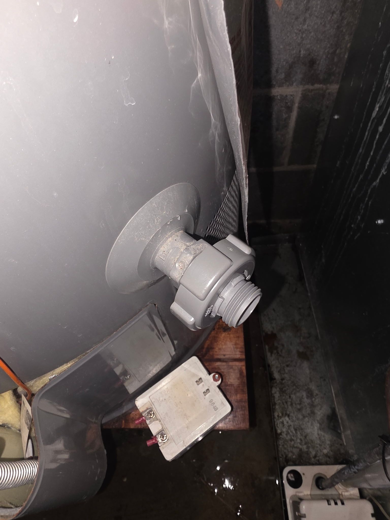 Damaged gray pipe fitting dripping water into a dark, wet floor area