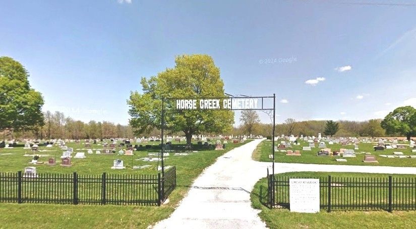 Entrance to a cemetery with a white path, black gate, and rows of grave markers under a blue sky
