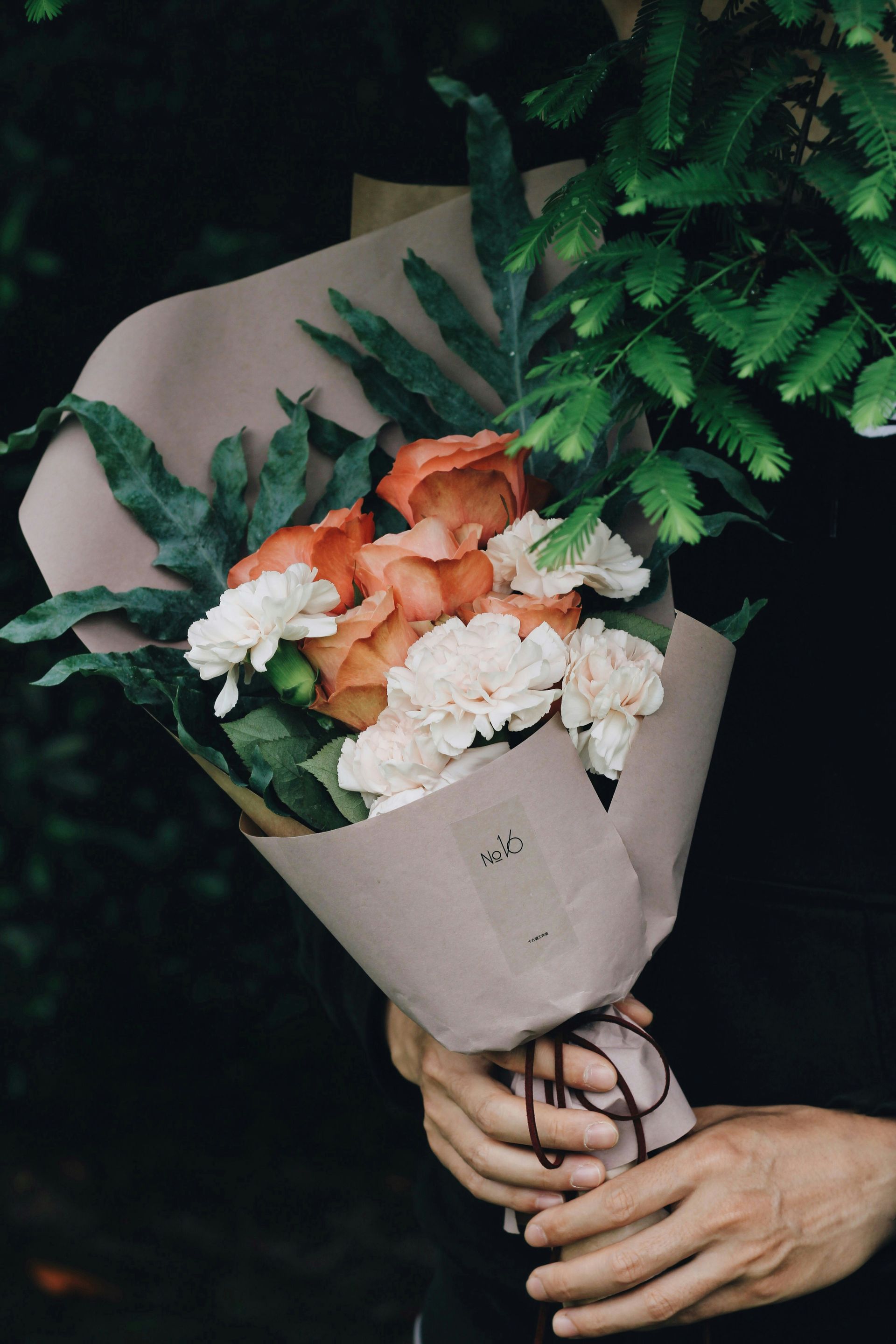 Hands holding a bouquet of pink and white flowers wrapped in beige paper against dark greenery