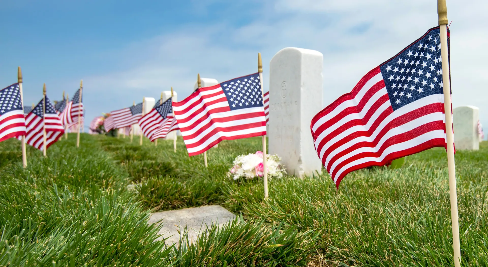 American flags waving in a grassy field near a white monument on a cloudy day