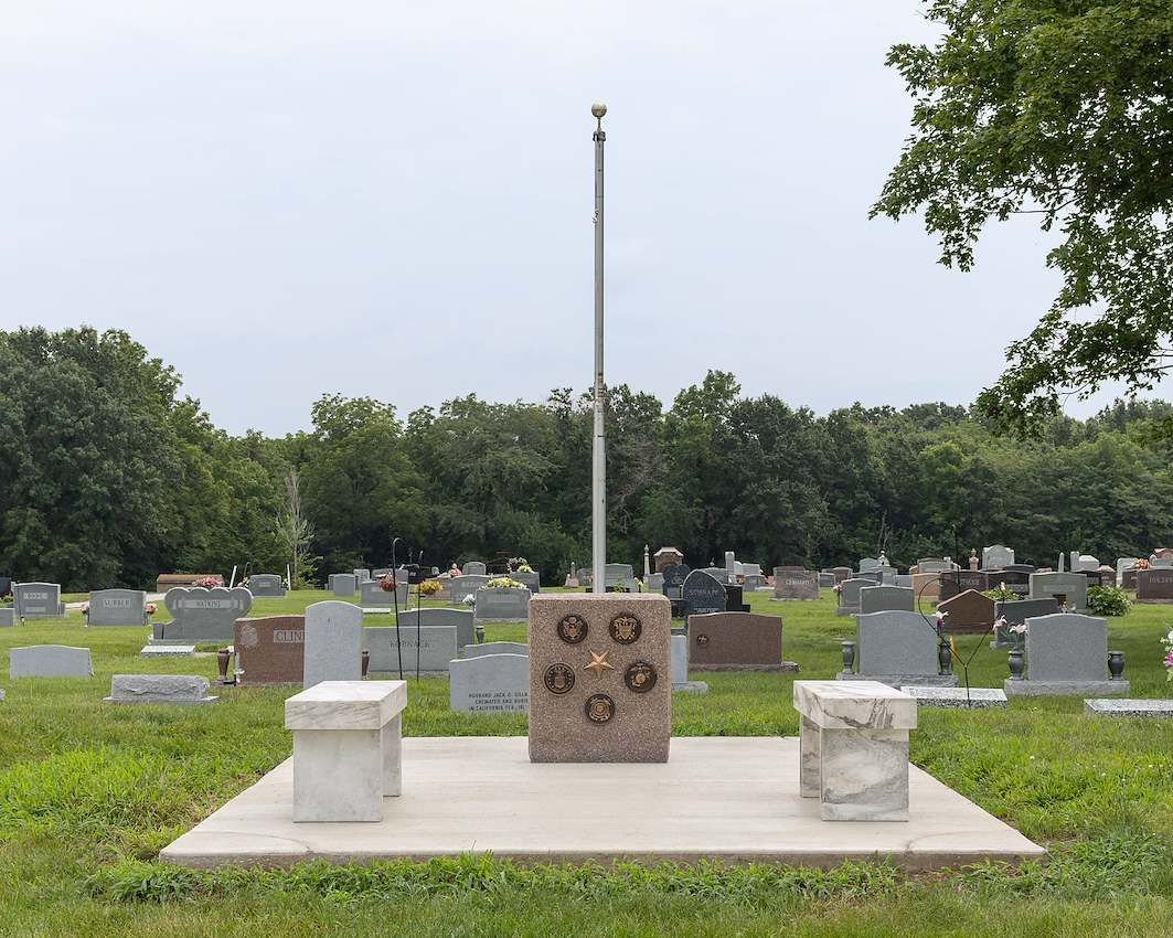 Memorial platform with flagpole in a grassy cemetery, surrounded by rows of gravestones.