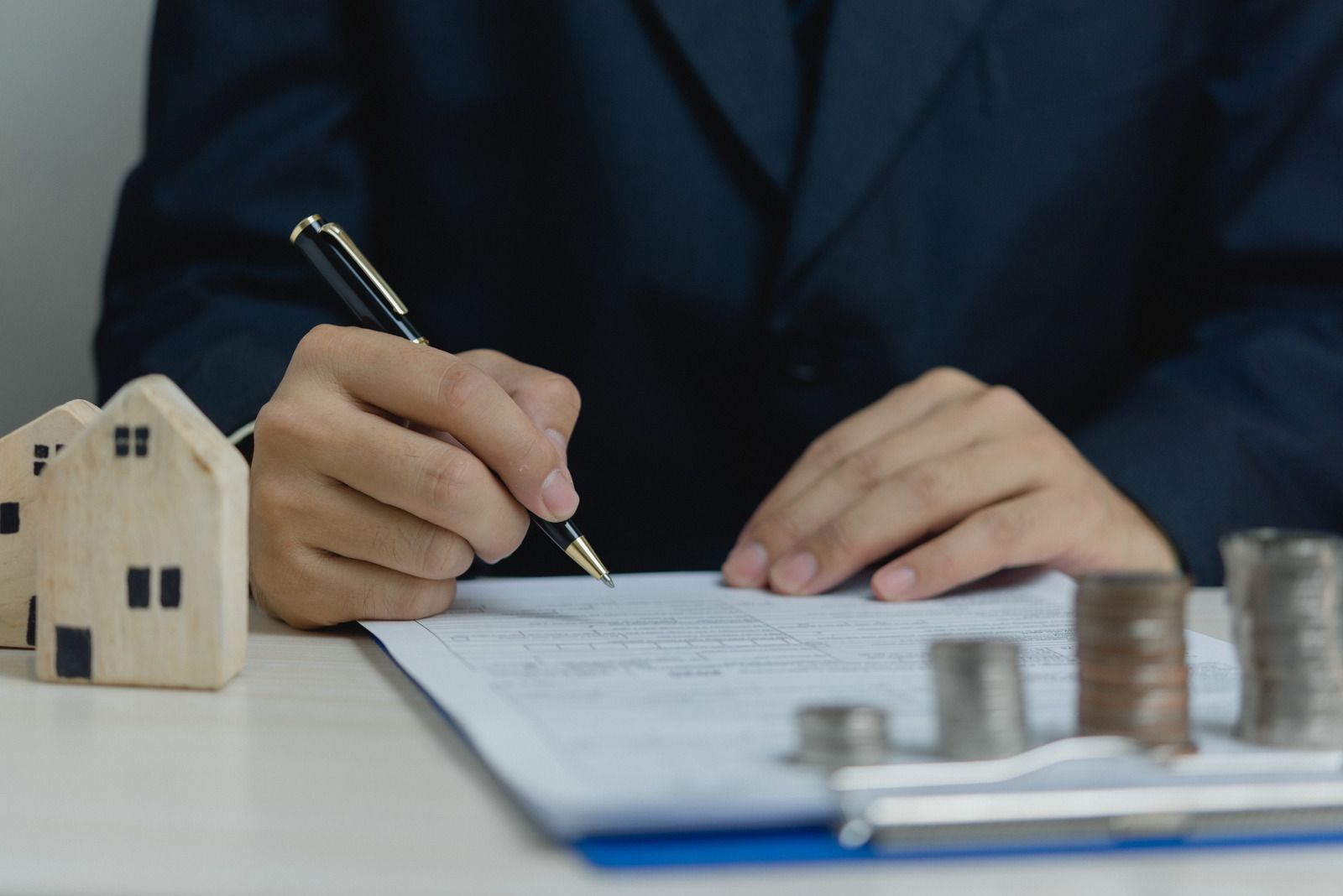 Person signing a document with miniature houses and stacks of coins on a table.