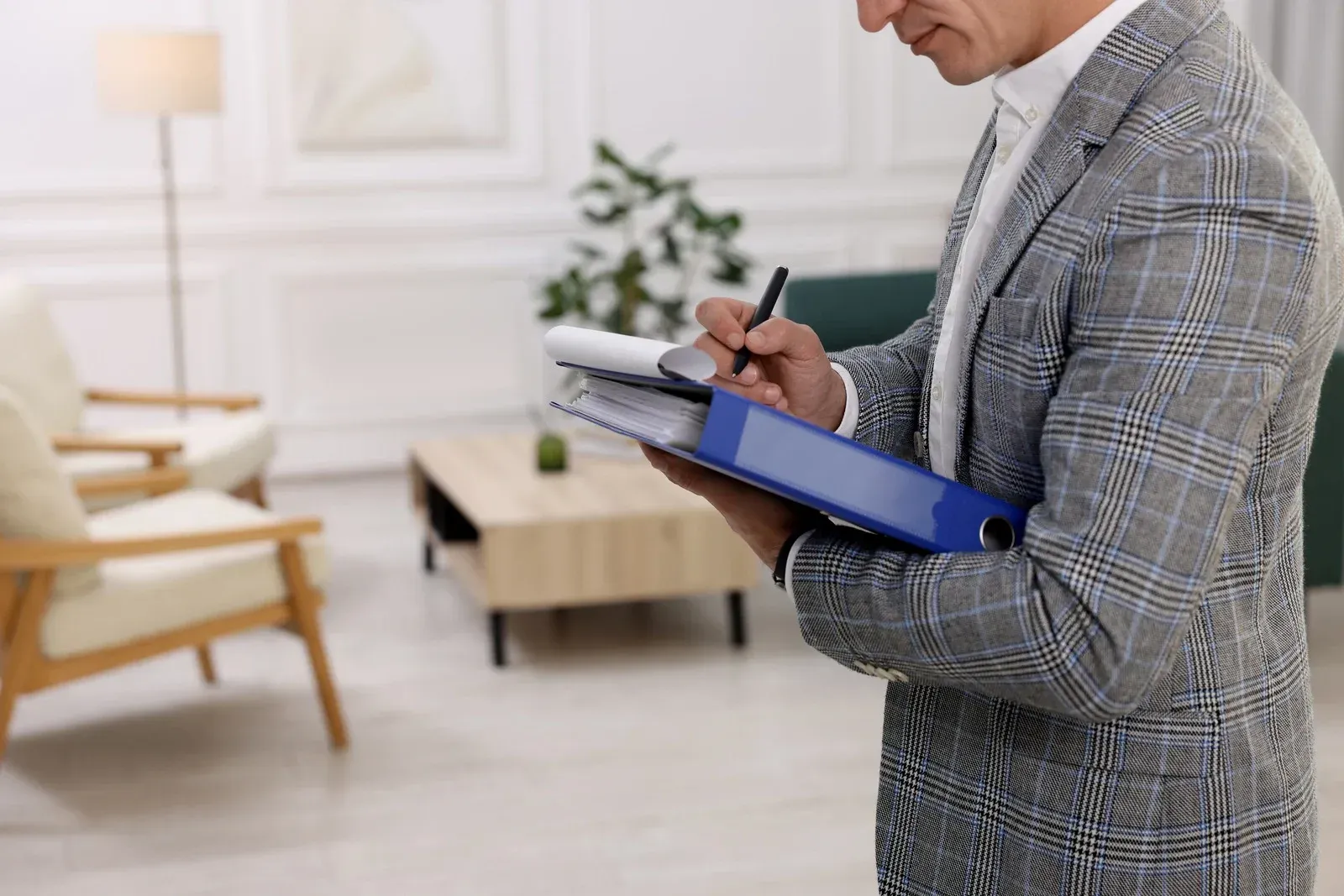 Man in a blazer writing in a blue binder, reviewing documents in a living room with furniture.