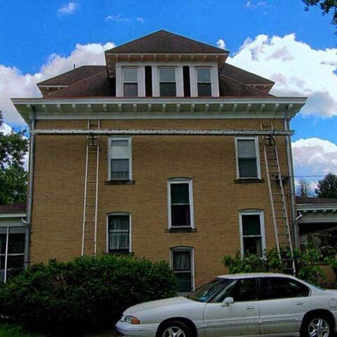 Two-story tan house with white trim and a white car parked in front under a blue sky