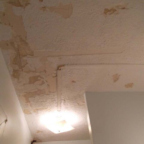Peeling, water-stained ceiling with a lit light fixture and a white wall corner