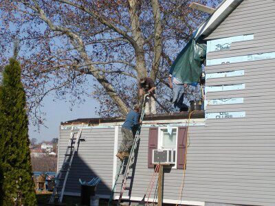 Person on a ladder repairing the roof of a gray house beside a tree