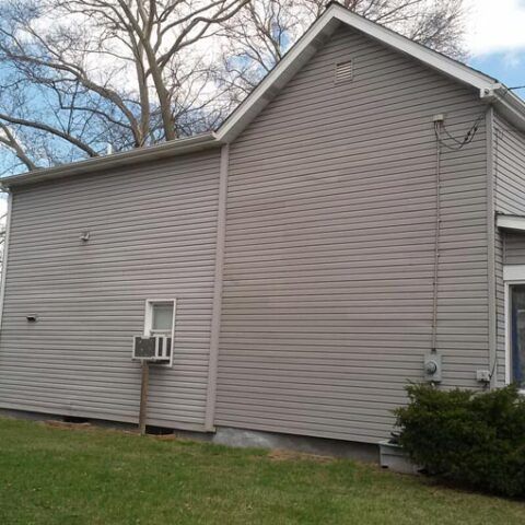 Side view of a beige house with vinyl siding, a small window AC unit, and a grassy yard.