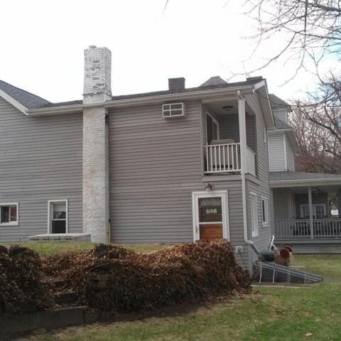 Gray two-story house with white trim, chimney, balcony, and a front porch surrounded by bushes.