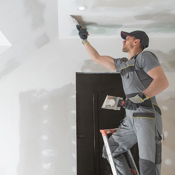 Painter on a ladder scraping and patching a white ceiling near a dark doorway.