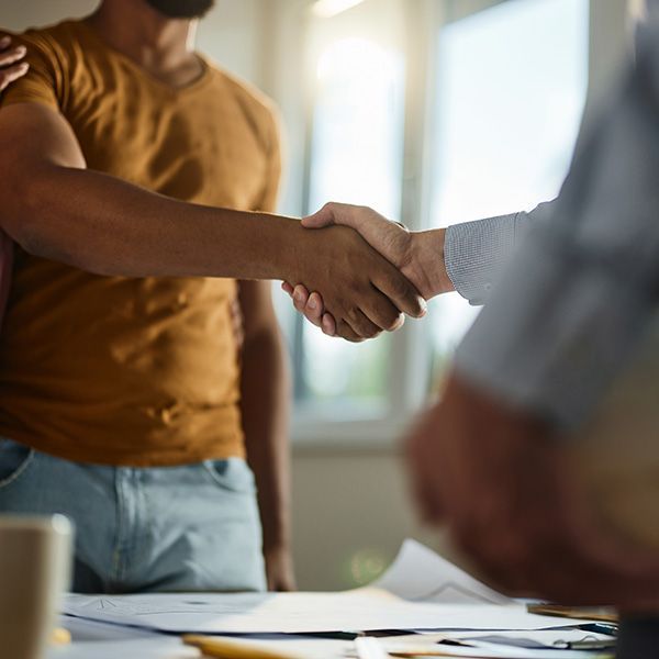 Two people shaking hands over a desk with papers, in a bright office setting