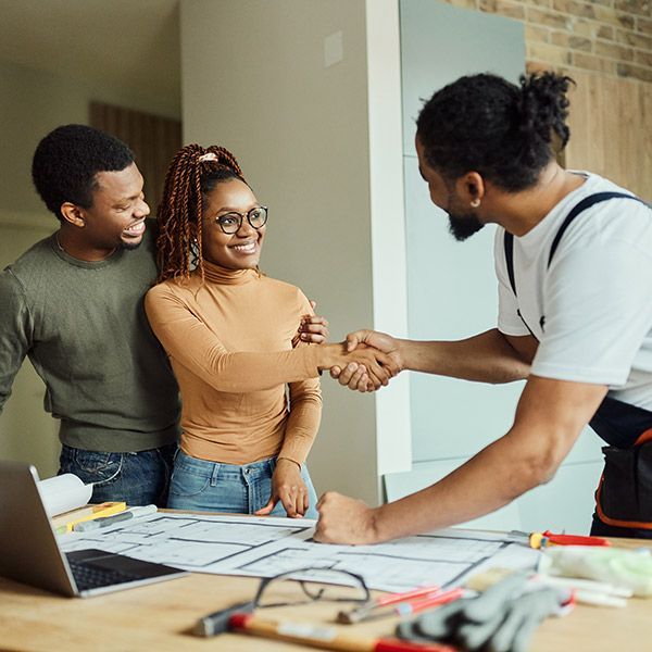 Three coworkers shake hands over a desk in a bright office, with a laptop and tools in the foreground.