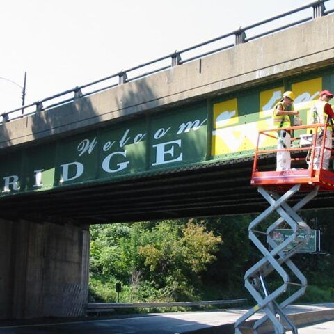 Workers on a scissor lift painting a green bridge overpass with “Welcome” text.