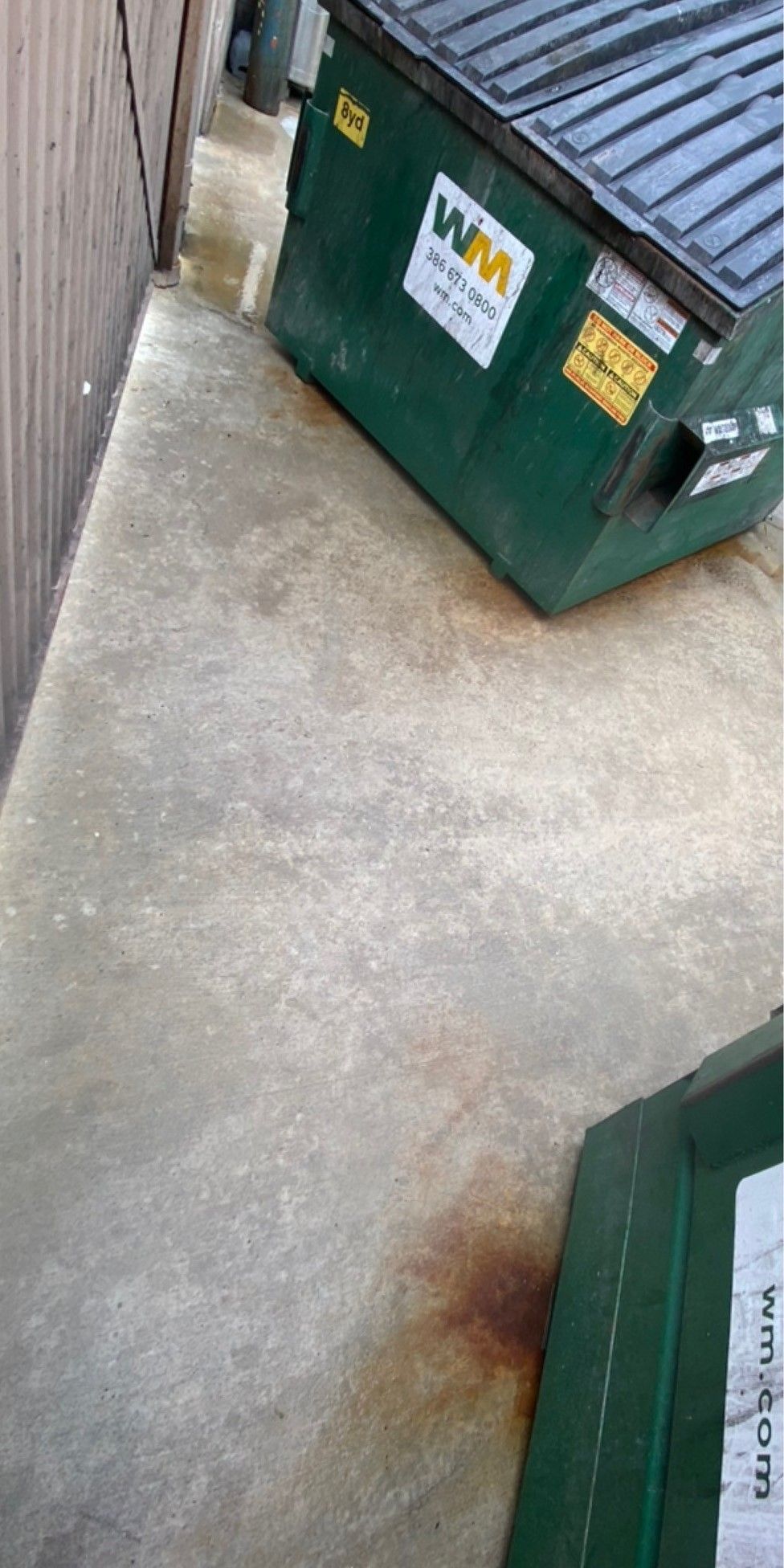Concrete alley with a green WM dumpster and a smaller bin, plus a rust stain on the ground