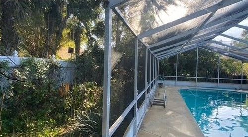 Covered outdoor pool beside a screened patio, with palm trees and greenery in the background.