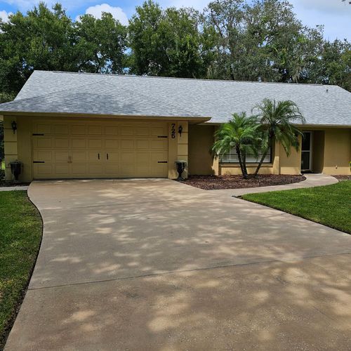 Single-story beige house with two-car garage, driveway, and front yard trees under a wavy metal roof