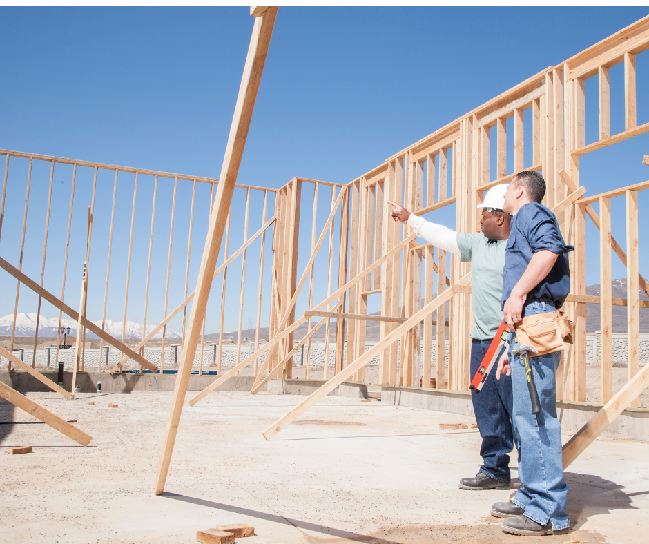 Two construction workers at a house frame; one points, the other watches. Blue sky and mountains in background.