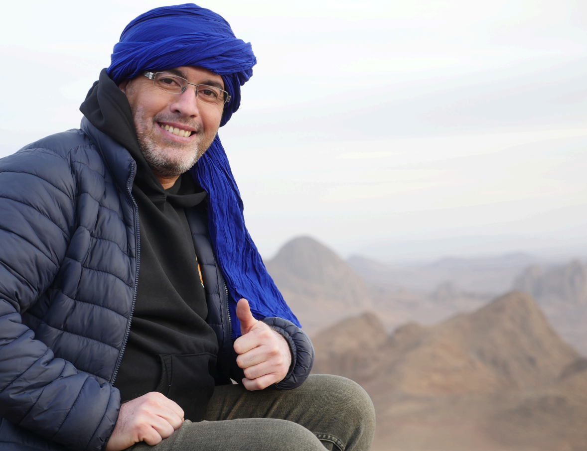 A smiling person wearing a blue turban and puffer jacket gives a thumbs-up against a backdrop of desert mountains.