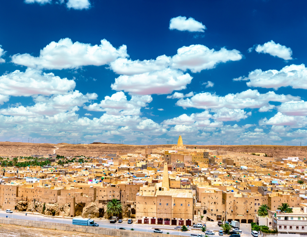 A panoramic view of Ghardaïa, Algeria, featuring traditional ochre-colored stone buildings under a bright, cloudy sky.