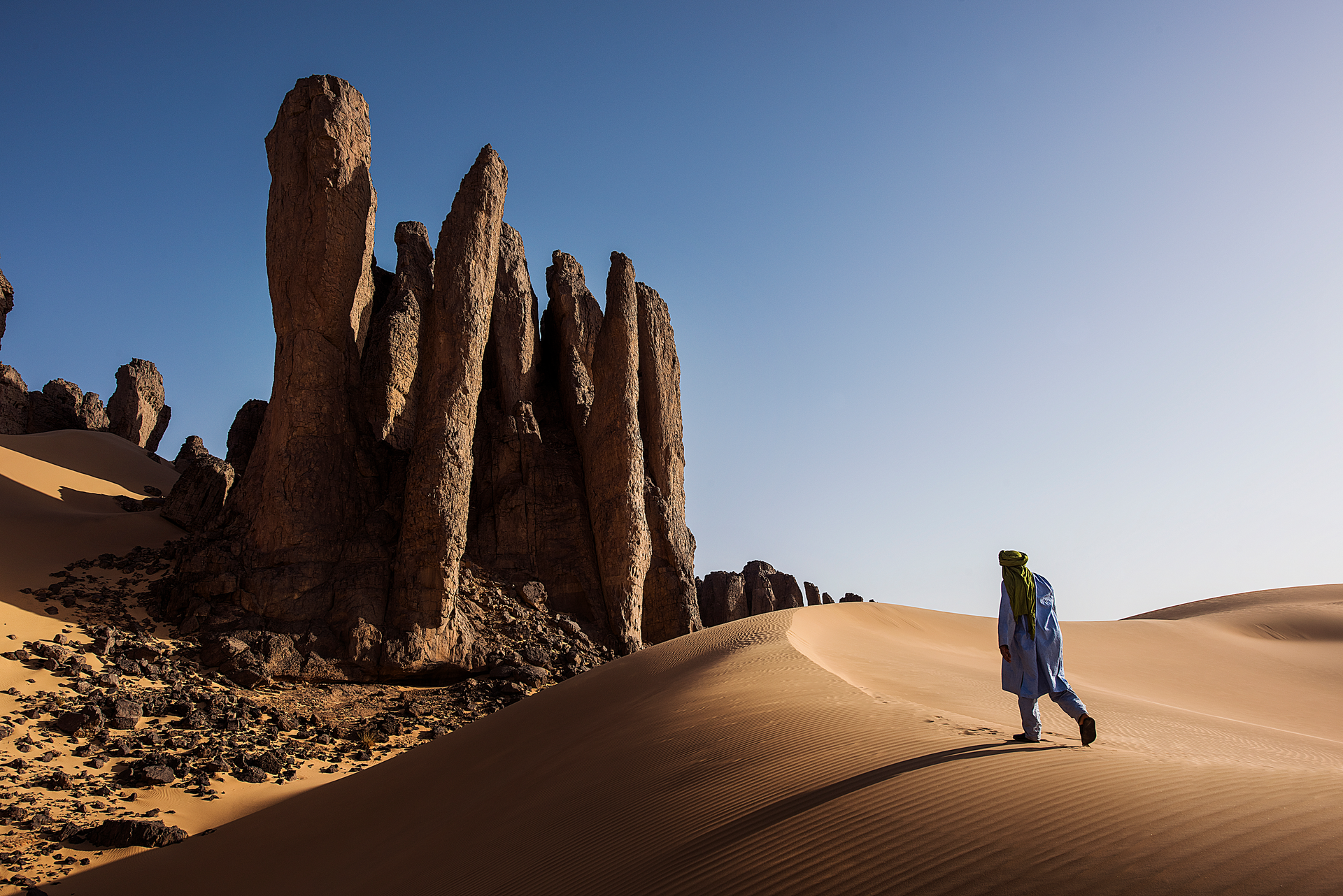 A person wearing traditional blue desert clothing walks across a sandy dune beside towering, weathered rock formations.