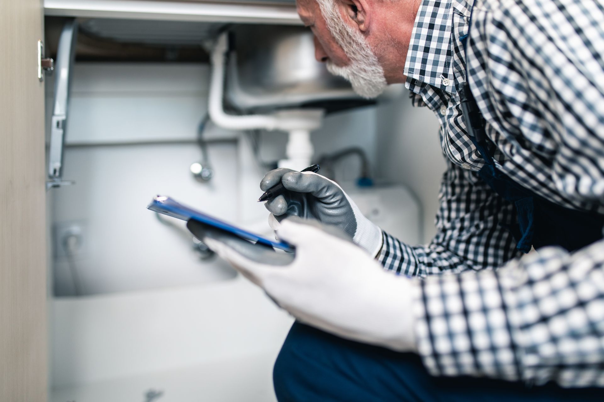 Plumber inspecting under sink pipes while taking notes on a tablet.