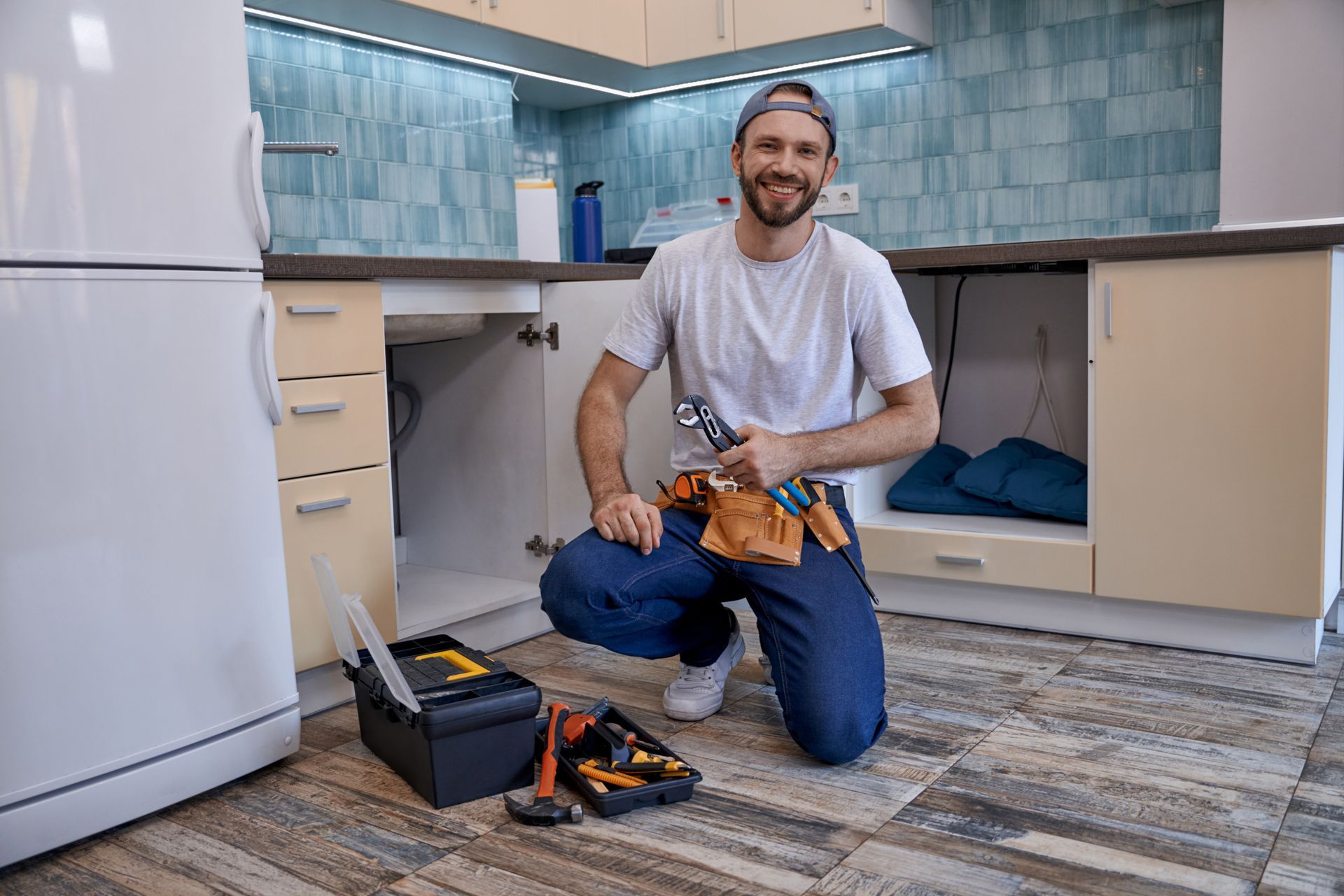 Home repair technician kneeling in kitchen beside open cabinets and toolkit.
