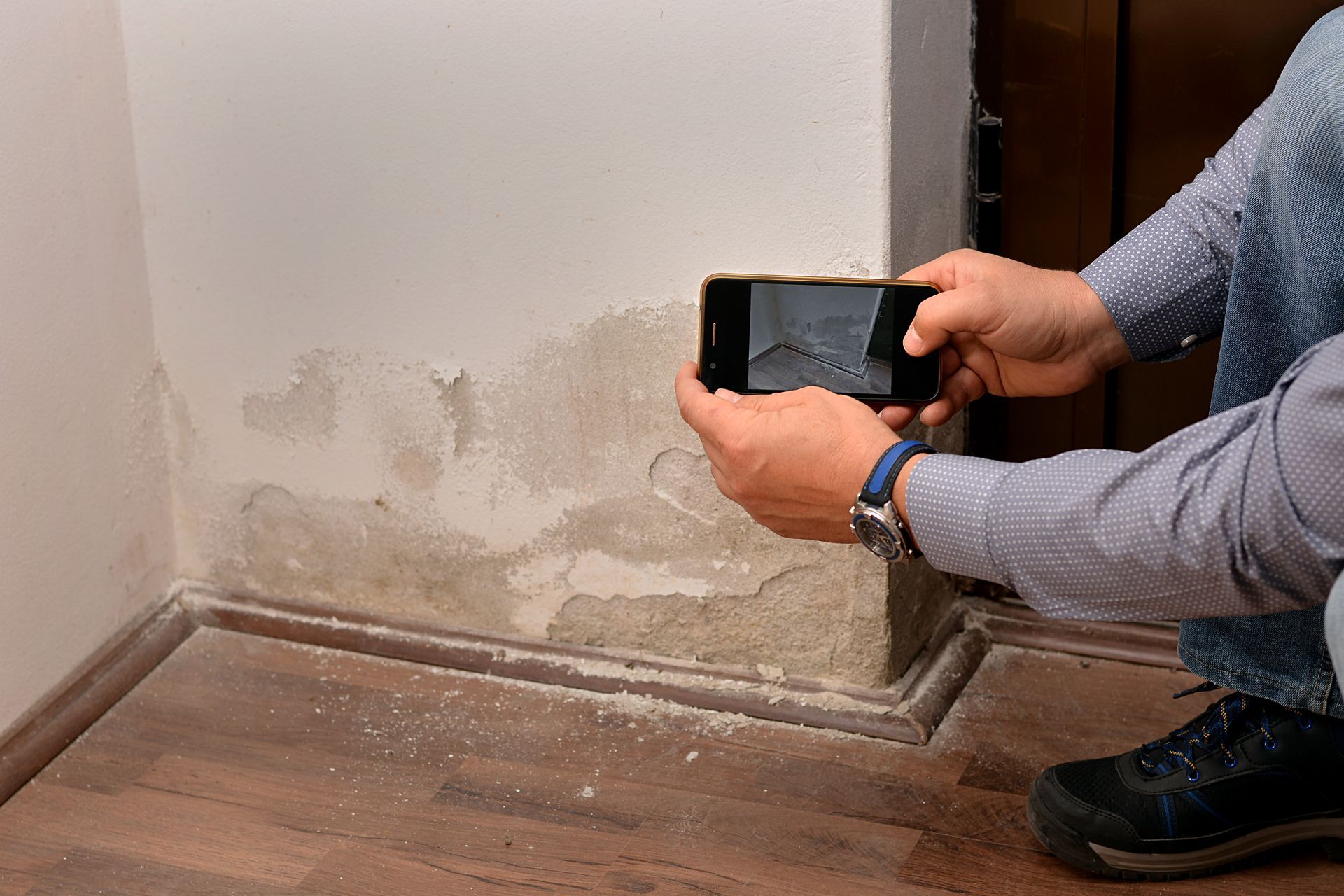 Person photographing water-damaged wall with peeling paint near floor molding.