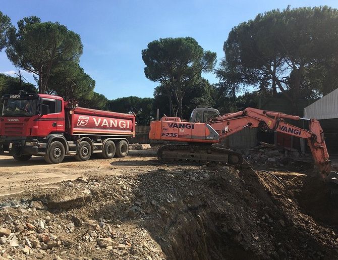 Un camion ribaltabile rosso e un escavatore arancione in un cantiere edile con alberi sullo sfondo.