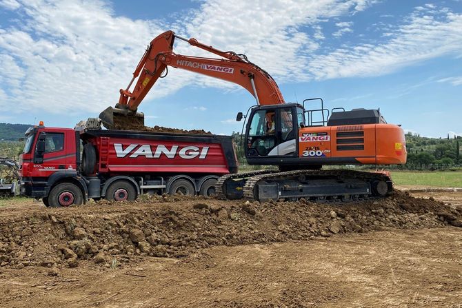 Un escavatore arancione carica terra su un camion ribaltabile VANGI rosso in un campo fangoso sotto un cielo nuvoloso.