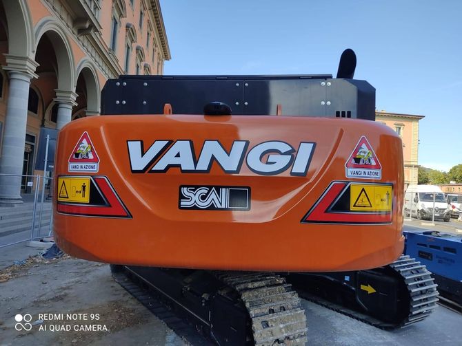 Escavatore cingolato VANGI arancione parcheggiato accanto a un edificio, vista posteriore con segnali di avvertimento e cingoli neri