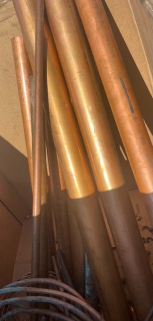 Copper-colored pipes stacked in a dim industrial space, with a coiled cable at the bottom left