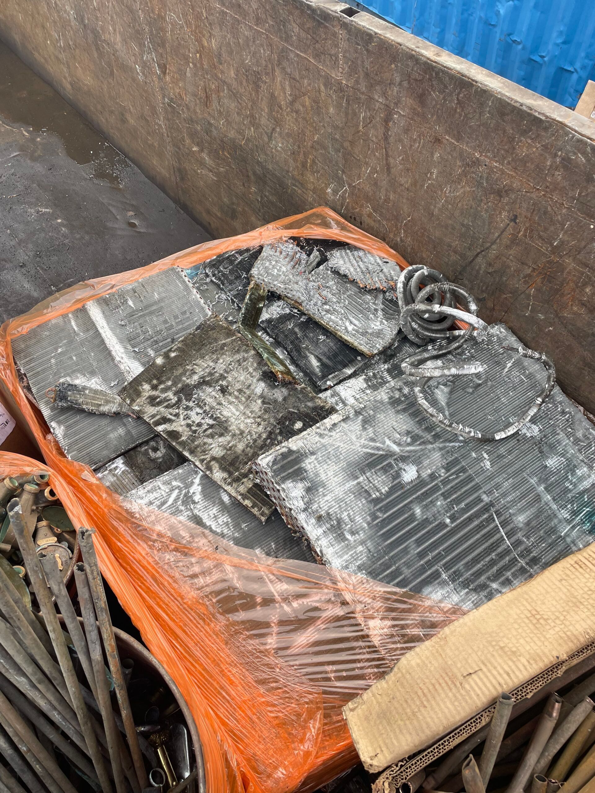 Orange-netted pallet of damaged gray metal parts in a concrete industrial area