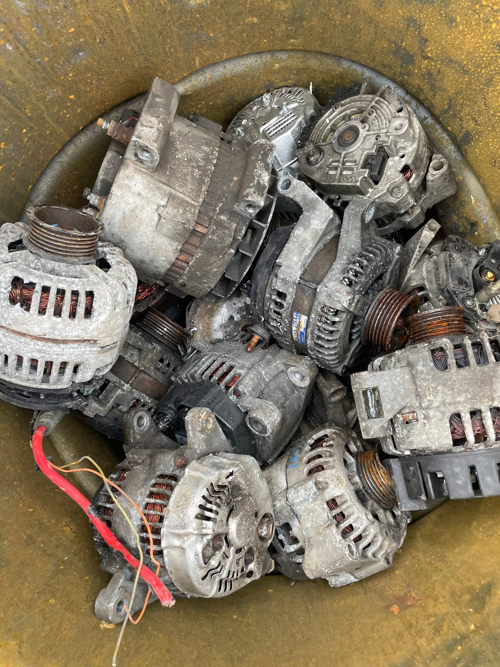 Pile of dirty used car alternators and metal parts in a yellow bin, with red wires visible