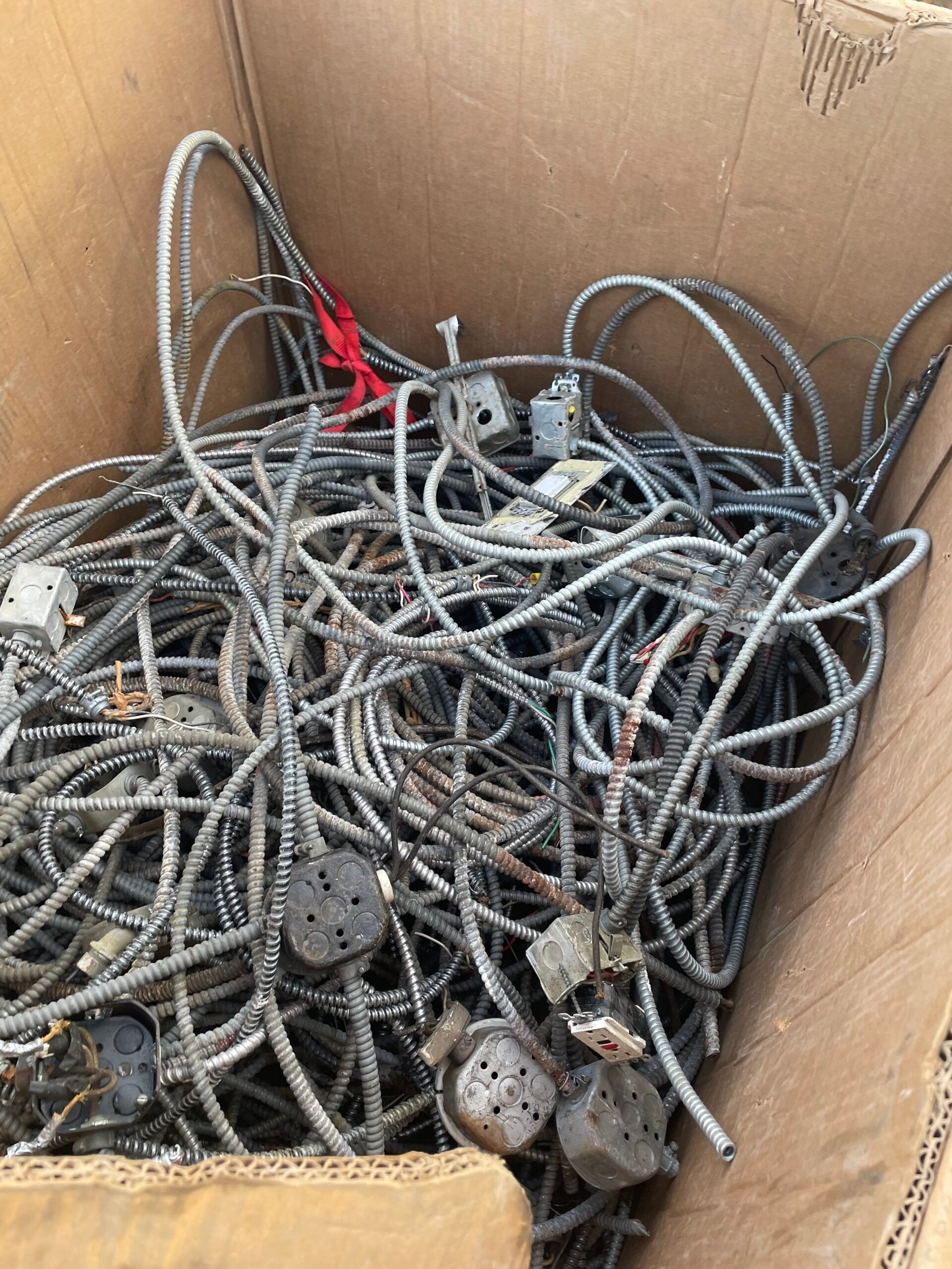 Cardboard box filled with tangled gray cords and cables, with a few black plugs visible.