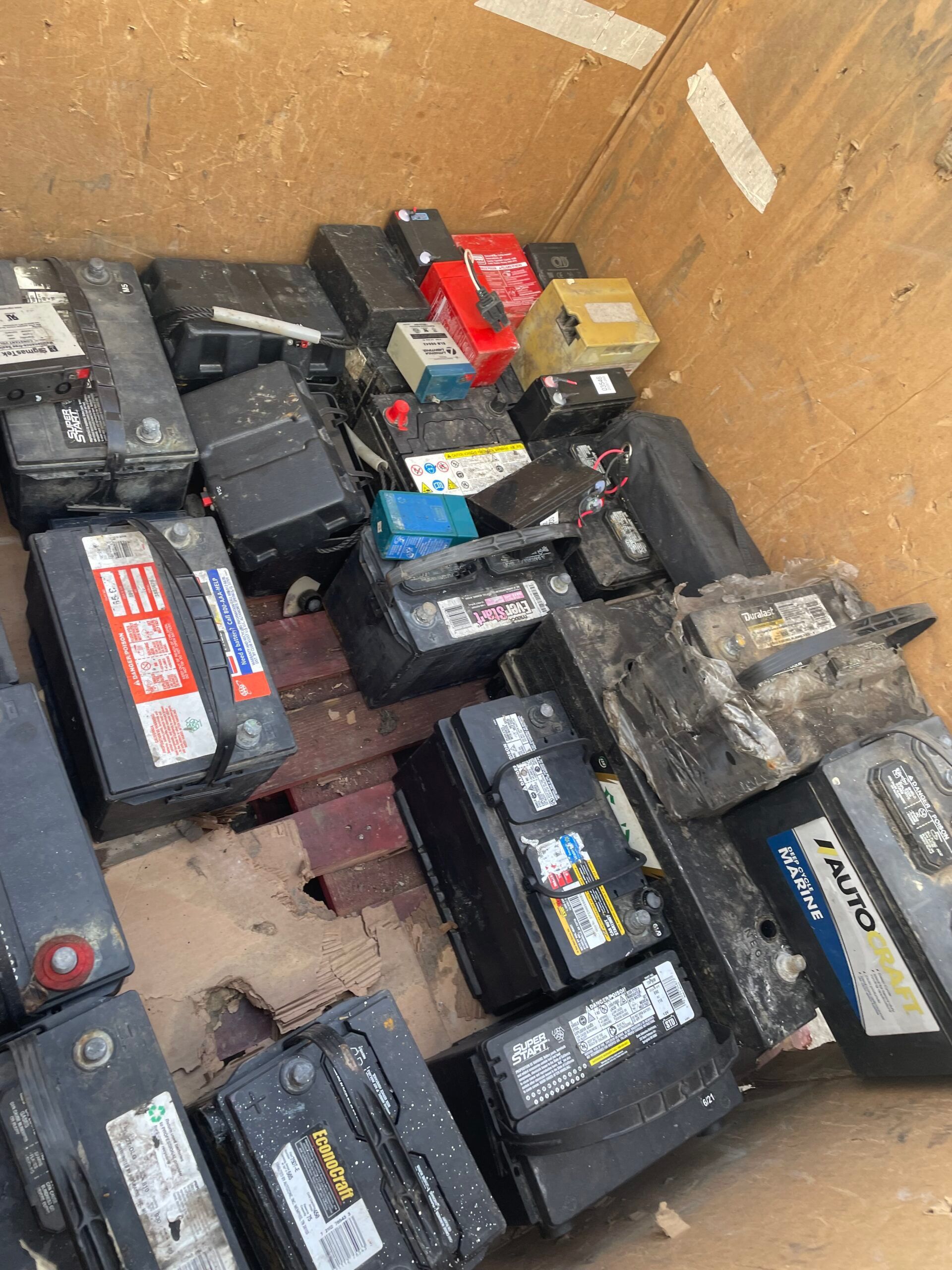 Scattered old car batteries in a cardboard box, including black, red, and yellow units.