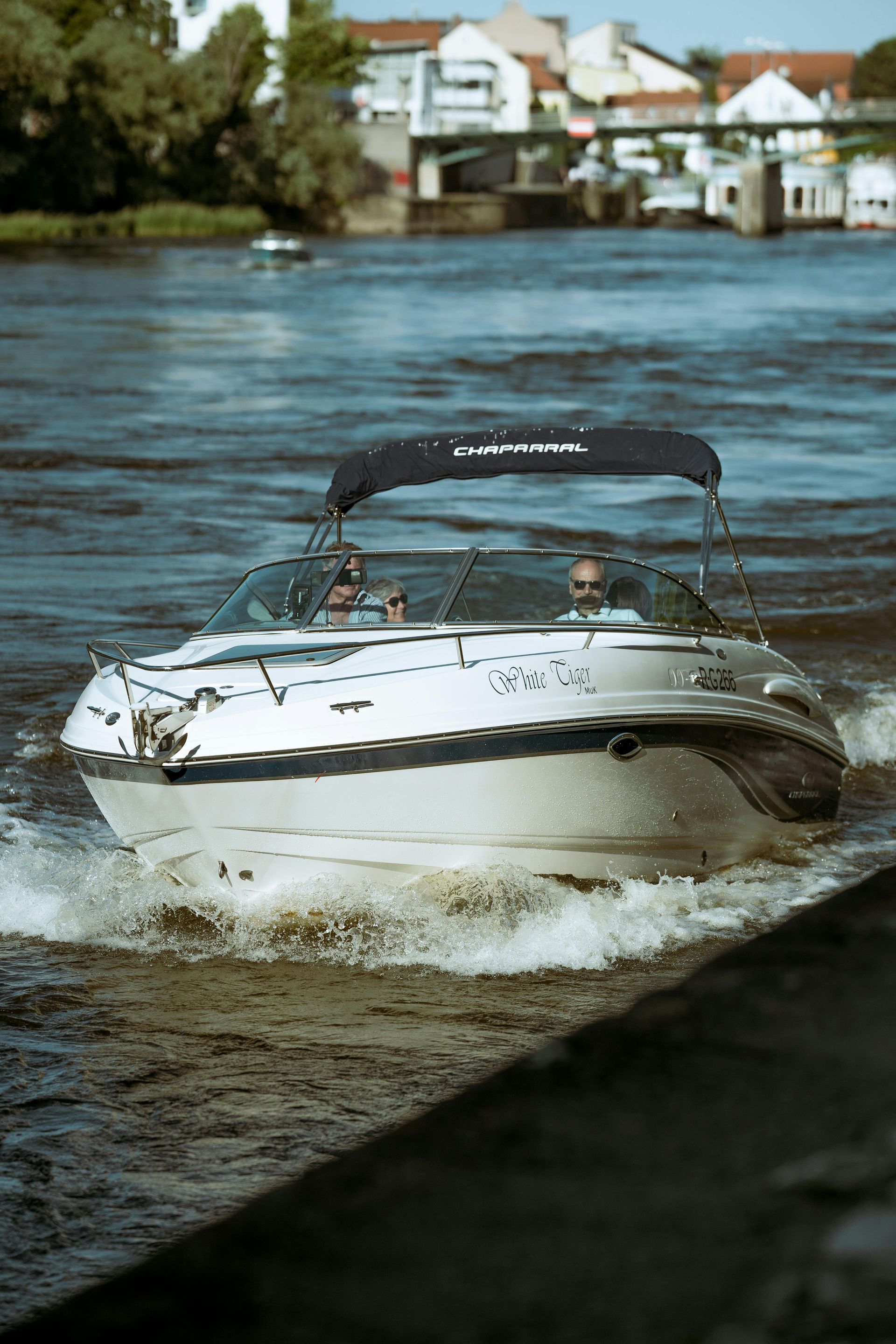 Motorboat on a river, two people visible.  White boat with black canopy. Buildings in the background.